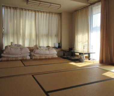 Sunlit tatami room with folded futons, low table and sheer curtains at Funayado Akitaya in Kanagawa