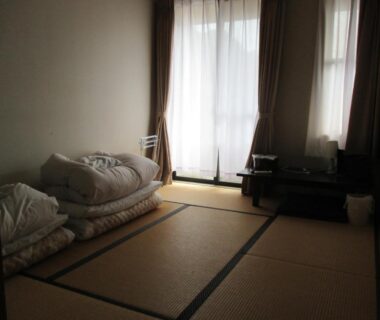 Simple tatami room with stacked futon bedding, low table and natural light from curtains at Funayado Akitaya Kanagawa
