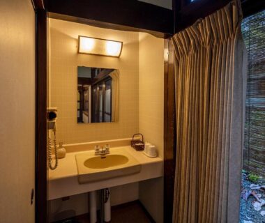 Compact wooden-framed washbasin with mirror, hairdryer, curtain and bamboo screen overlooking small garden at Furusato ryokan in Gifu