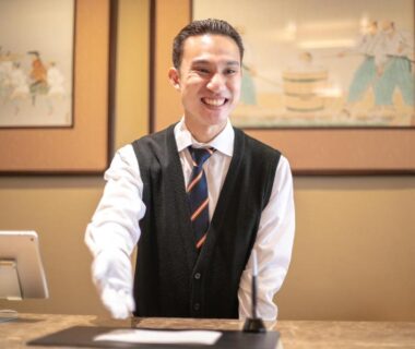 Smiling receptionist at Furuya Ryokan front desk with traditional framed artwork, Shizuoka, Chūbu, Japan