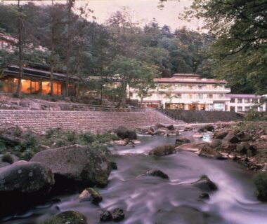 Dusk riverside scene at Gensenkan with lit ryokan buildings beside a rocky stream and forested banks in Tochigi