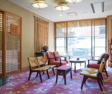 Sunlit lounge with wooden lattice screens, upholstered chairs, low tables and bookshelf at Gingetsu in Nagano