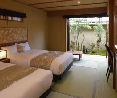 Tatami-floored twin room with two low beds, wooden desk, lantern lamp and small garden view through sliding door