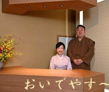Two ryokan staff in kimono behind wooden reception desk with flower arrangement and welcome sign at Gion Oyado Kikutani Kyoto