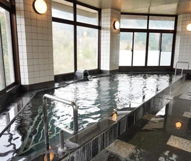 Tiled indoor onsen pool with metal handrails and large windows overlooking green hillside