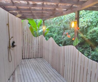 Covered wooden deck with outdoor shower, lanterns and slatted fence beside green foliage at Guest House Churaumi Shizuoka