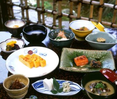 Assortment of small Japanese dishes including tamagoyaki, grilled fish and miso soup on tray near bamboo screen