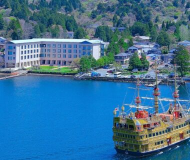 Sightseeing pirate-style boat on blue lake beside Hakone Hotel waterfront with tree-covered hills and shoreline town in Kanagawa