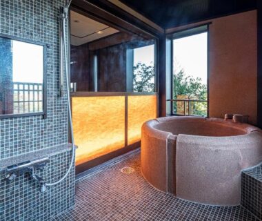 Circular tiled soaking tub with shower area and large windows overlooking trees at Hakone Kyuan