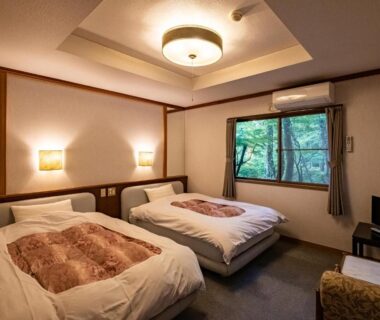 Two single beds with bedside lamps, window revealing garden trees, compact ryokan room interior in Hakone, Kanagawa