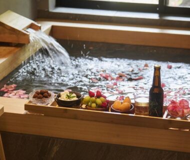 Wooden soaking tub with flowing spout, fruit and snacks on tray and rose petals by window at Hakone Retreat
