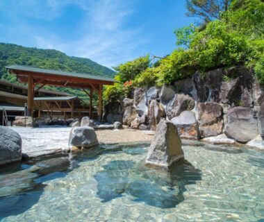 Stone-lined outdoor onsen pool with clear water, rock formations and green mountain backdrop at Hakonenomori Okada, Kanagawa