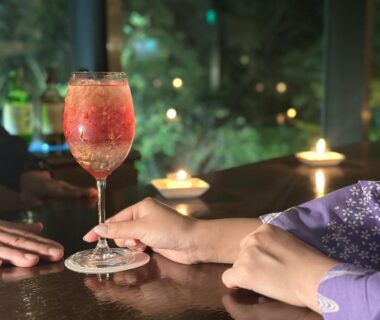 Stemmed glass with pink drink on wooden bar, hands in kimono sleeve, candlelight at Hana to Hana, Tochigi Japan