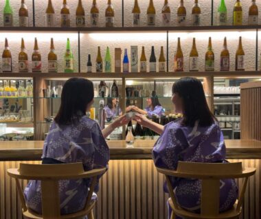 Two guests in yukata clinking glasses at a wooden bar with illuminated sake bottles, Hana to Hana, Tochigi, Kantō, Japan