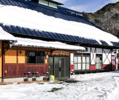 Snow on roof and ground outside Hanasaku Onsen Yoshiyaso entrance with chairs and mountain backdrop in Gunma