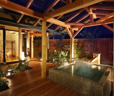 Stone bath on wooden deck under timber roof with warm lantern lighting and garden planting at Hanashibuki, Chiba
