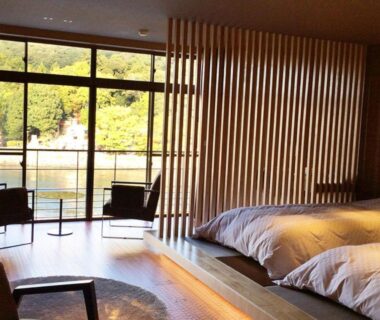 Tatami room with futon bedding, wooden slatted divider and riverside balcony overlooking trees at Hanayashiki Ukifune-en, Kyoto