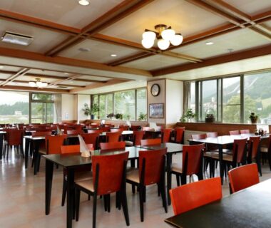 Spacious ryokan dining room with wooden ceiling, rows of tables and red chairs, large windows facing hills