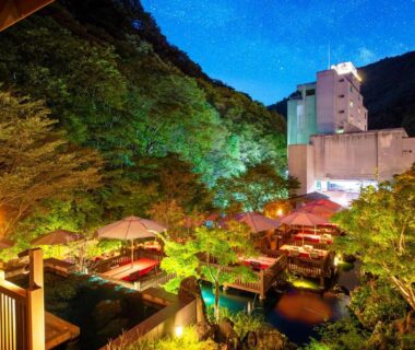 Illuminated terraced outdoor baths among trees with mountain backdrop at Harataki in Fukushima