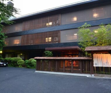 Traditional wooden ryokan facade with noren curtain, warmly lit entrance and small landscaped trees at Hatago Kintoen in Kanagawa
