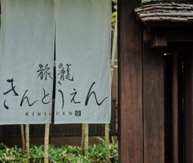 White noren with calligraphy hanging at a wooden ryokan entrance, greenery visible, Hatago Kintoen in Kanagawa