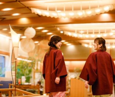 Two guests in yukata walking through warm wooden ryokan lobby with seating and soft lighting