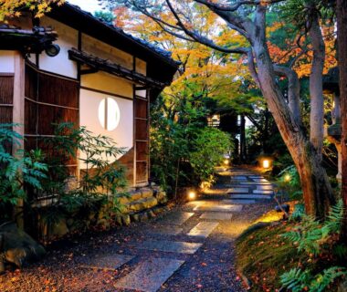 Stone garden pathway lit by lanterns leading to a wooden teahouse entrance with shoji window and autumn foliage