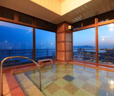 Indoor tiled onsen bath with floor-to-ceiling windows overlooking coastal harbor and distant shoreline
