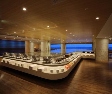 Spacious dining hall with central serving counter, wooden ceiling and floor-to-ceiling windows with blue horizon view