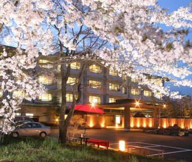 Night view of lit ryokan entrance with blooming cherry trees and parked cars, Hibikino, Gunma, Kantō