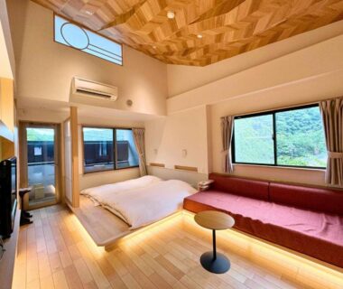 Bright Japanese-style room with low futon bedding, wooden floors, geometric ceiling, long bench and windows overlooking trees