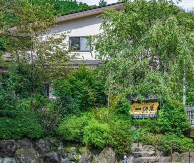 White ryokan building behind lush foliage by stone riverbank with wooden signboard at Hidamari Ichinoyu, Kanagawa