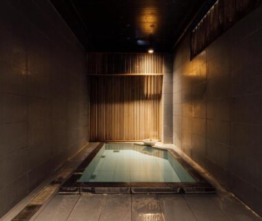 Rectangular indoor onsen with stone floor, wooden slatted wall and ambient lighting at Hinatamikan in Gunma
