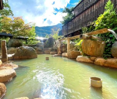 Rock-lined outdoor hot spring pool with bamboo spouts and mountain backdrop at Hirayukan Gifu