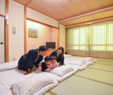 Family in yukata on futon bedding in tatami room with curtained window and low wooden ceiling