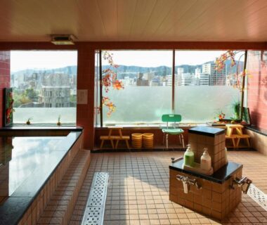 Tiled indoor onsen with stools, wash stations and floor to ceiling windows overlooking Hiroshima city and hills