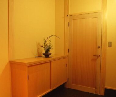 Simple wooden entrance with cabinet, floral arrangement, wooden door and warm lighting at Hitomaru Kadan in Hyōgo, Kansai
