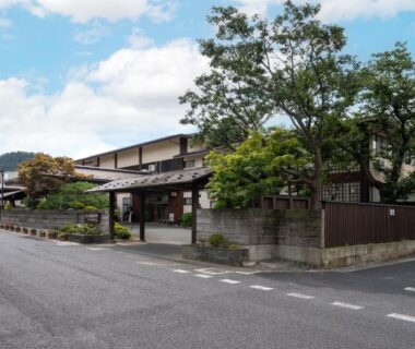 Street-facing exterior of Hohoemino Kuyufu Tsuruya with traditional wooden facade, entrance gate and surrounding greenery in Yamagata