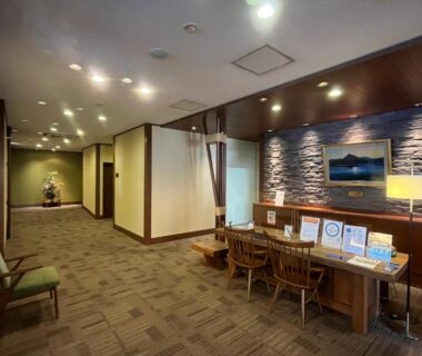 Carpeted ryokan lobby with reception desk, brochure table, wooden chairs and corridor lighting in Hokkaidō