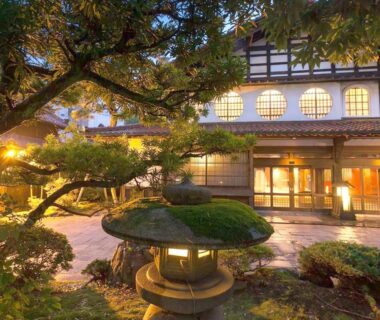 Stone lantern and manicured pine framing lit traditional ryokan entrance with shoji windows in Ishikawa