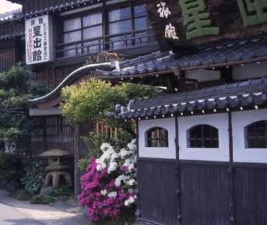 Traditional wooden ryokan exterior with tiled roof, shoji windows, stone lantern and blooming azaleas at Hoshidekan entrance in Mie