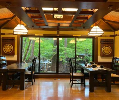 Wooden ryokan dining room with tables, pendant lights and leafy garden visible through large glass doors