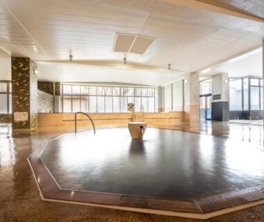Spacious communal indoor onsen with central octagonal bath, tiled floor, handrail and windows at Hotel Daiheigen in Hokkaidō