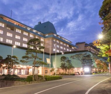 Illuminated multi-story ryokan facade with entrance canopy, landscaped pines and street in front at dusk in Iwate