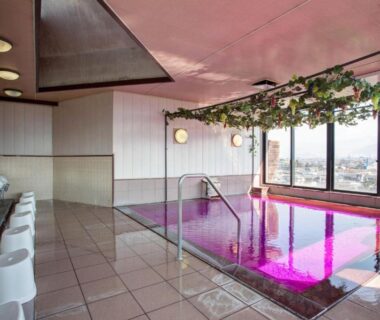 Indoor onsen bath with magenta lit water, tiled floor, washing stools and large windows overlooking Yamanashi city