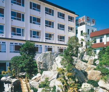 Hotel Heian facade with rock garden, wooden bridge and landscaped trees under blue sky in Yamanashi