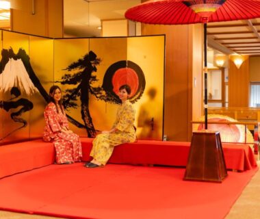 Two women in kimono seated on red platform by gold folding screen, traditional umbrella and wooden interior