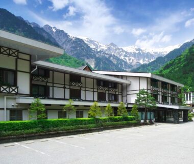 White ryokan building with balconies and mountain peaks in the background, paved forecourt and landscaped shrubs