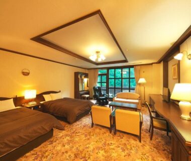 Carpeted twin bedroom with two beds, seating area, desk and large window with outdoor greenery