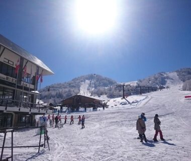 Skiers on a snowy slope beside a lodge under bright sun at Hotel Ichibokaku in Nagano mountains